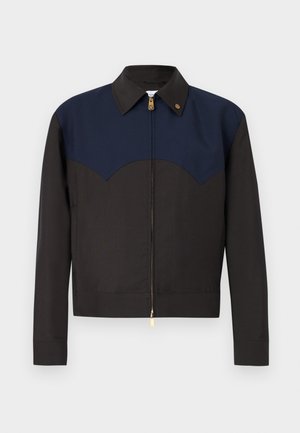 Mand stående iført sort og marineblå jakke, grøn mønstret skjorte, sorte bukser, sorte støvler, med hænderne i lommerne mod ensfarvet grå baggrund.