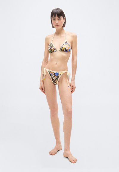 Λεπτή γυναίκα που στέκεται ξυπόλυτη φορώντας πολύχρωμο μπικίνι με δεσίματα στο πλάι, με λουλουδάτα και αφηρημένα σχέδια πάνω σε απλό ανοιχτό φόντο.