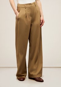 Pantaloni a gamba larga in tessuto beige, con pieghe frontali, tasche inclinate e chiusura con un bottone in vita. Texture liscia.