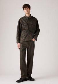 Mladenič stoji v temno rjavem jeans jakni in ujemajočih širokih hlačah, črnih čevljih in srebrni zapestnici na nevtralni podlagi.