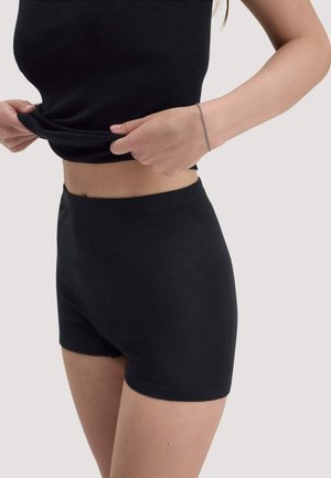 Femme portant un short noir taille haute et un top noir sans manches, soulevant légèrement le top pour dévoiler le ventre, avec un bracelet au poignet.