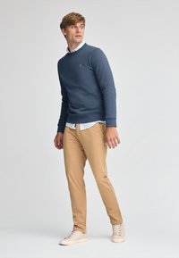 Marinblå sweatshirt med rund halsringning, över en ljusblå skjorta med krage, i kombination med tanfärgade chinos och ljusbeige sneakers. Enkelt, avslappnat design.
