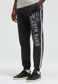 Schwarze Jogginghose mit roten Kordeln, seitlichem wei ßen Streifen und großem grauem Grafikdruck am Bein. Hergestellt aus weichem, strukturiertem Stoff.