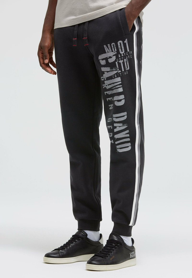 Schwarze Jogginghose mit roten Kordeln, seitlichem weißen Streifen und großem grauem Grafikdruck am Bein. Hergestellt aus weichem, strukturiertem Stoff.