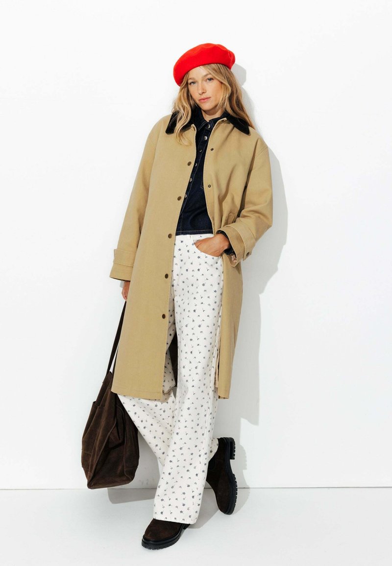 Manteau long beige avec un col noir, chemise marine, pantalon floral blanc, bottes marron et béret rouge. Un sac à bandoulière marron complète la tenue.