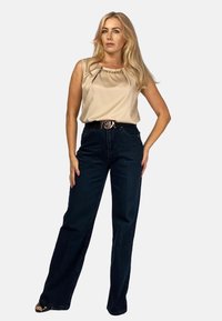 Beżowy satynowy top na ramiączkach z plisowanym wzorem i złotym detalem w postaci łańcuszka, w połączeniu z ciemnoniebieskimi dżinsami o wysokim stanie oraz czarnym paskiem ze złotą klamrą.