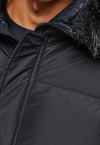 Detail osoby v černé prošívané bundě s kapucí lemovanou kožešinou a částečně rozepnutým předním zipem.