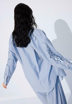 Vrouw met lang donker haar, gekleed in een lichtblauw gestreept shirt en broek met bloemenborduursels op de mouwen, leunend tegen een witte muur.
