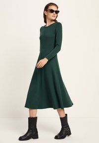 Frau trägt ein dunkelgrünes langärmliges Midikleid, schwarze Stiefel bis zur Wade und schwarze Sonnenbrille, steht vor einem einfarbigen hellen Hintergrund.