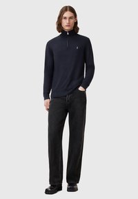 Marineblauer Strickpullover mit hohem Kragen und halben Reißverschluss, kombiniert mit lockeren schwarzen Jeans aus Denim und schwarzen Schuhen. Minimalistisches Logodesign.