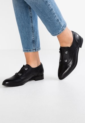 Osoba w czarnych skórzanych butach na niskim obcasie z srebrnymi guzikami, w połączeniu z podwiniętymi niebieskimi dżinsami, stoi na białej podłodze.