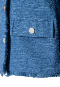 Veste bleue texturée avec un bas à franges, une poche avant avec fermeture à bouton et un subtil motif tissé sur tout le tissu.