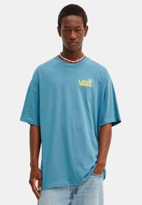 Camiseta azul de gran tamaño con escote redondeado y mangas cortas, con un logo amarillo de "VANS" en el pecho, combinada con unos vaqueros de mezclilla claros.