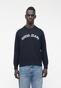 Tumši zils adīts džemperis ar apaļu kaklu, ribotu apakšu un baltu uzrakstu "GUESS JEANS" uz krūtīm. Kopā ar gaiši zilām džinsiem.