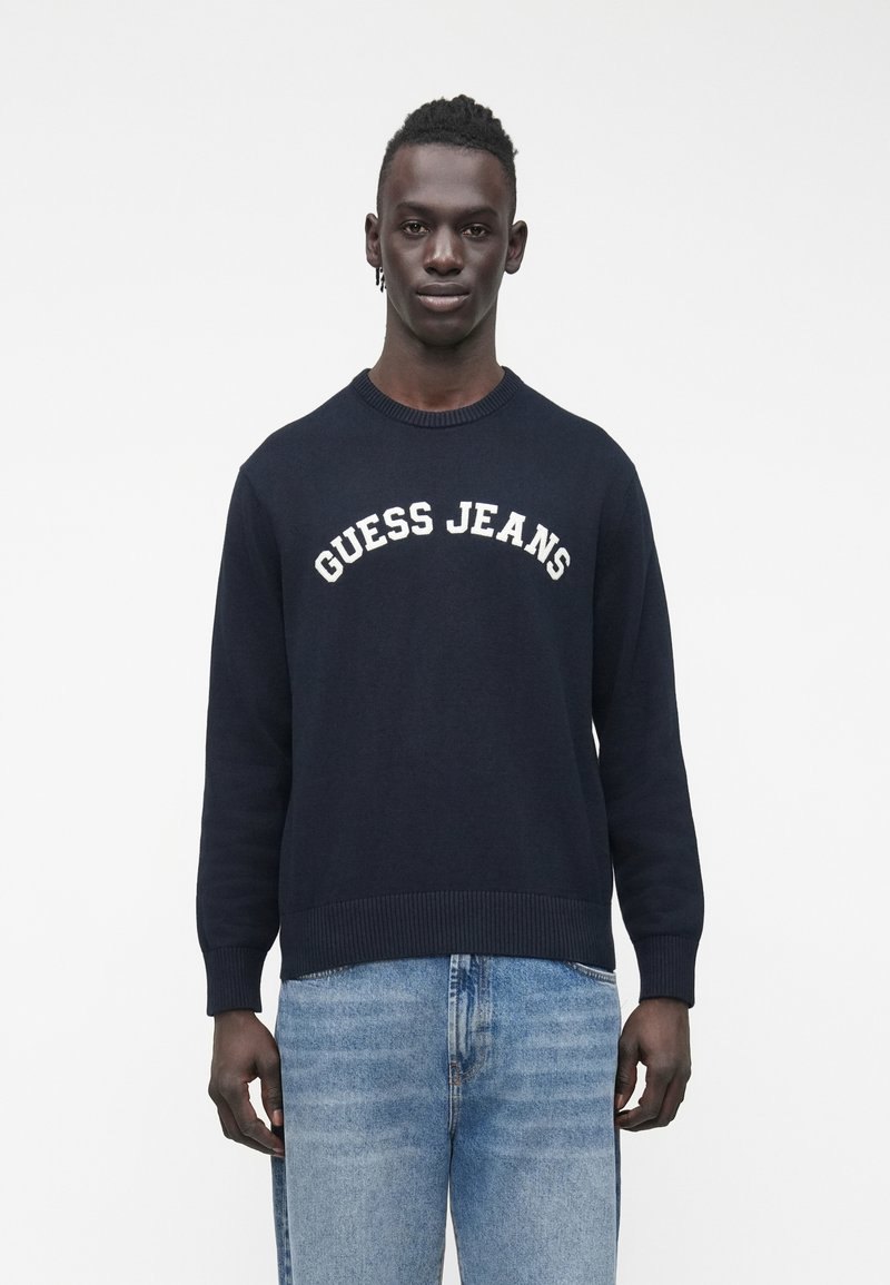 Tumši zils adīts džemperis ar apaļu kaklu, ribotu apakšu un baltu uzrakstu "GUESS JEANS" uz krūtīm. Kopā ar gaiši zilām džinsiem.