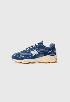 Blauer Wildleder New Balance 1000 Sneaker mit weißen Schnürsenkeln und beiger Sohle, seitlich gesehen auf einem einfarbigen hellgrauen Hintergrund.