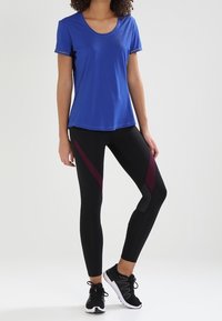 Blå sport t-shirt med korta ärmar, parat med svarta leggings med maroon och nätdetaljer, och svarta sneakers.