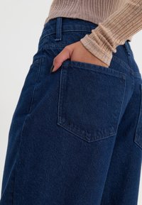 Mano con esmalte de uñas negro metida en el bolsillo trasero de unos pantalones vaqueros de mezclilla azul oscuro, llevando una camiseta de manga larga beige de canalé.