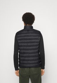 Person med kort lockigt hår som bär en svart vadderad väst över en svart långärmad tröja och mörkgröna byxor, vänd bort.