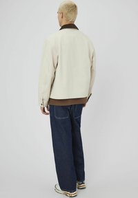 Veste beige avec un col sombre, associée à un haut marron et un jean large bleu foncé. La veste présente un design épuré et simple.