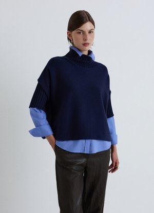 Donna con capelli pettinati all'indietro indossa un poncho blu navy sopra una camicia azzurro chiaro e pantaloni scuri, in piedi davanti a uno sfondo chiaro e uniforme.