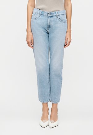 Φωτεινό μπλε τζιν ψηλόμεσο με ίσια μπατζάκια, φορεμένο με μπεζ αμάνικο τοπ και λευκές μυτερές γόβες, όρθιο μπροστά σε λευκό φόντο.