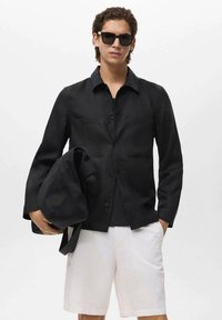 Sort skjorte med knapper, med to forlommer, båret over en sort top. Hvide shorts og en sort taske. Monokrom farveskema og enkelt design.