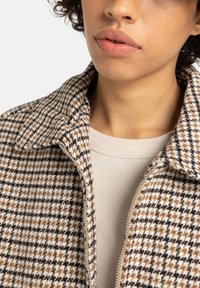 Beżowa i brązowa kurtka w wzór houndstooth z kołnierzem, zamkiem błyskawicznym oraz teksturowanym materiałem noszona na jasnobeżowej bluzce.