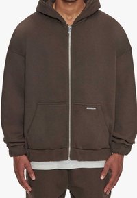 Brauner Kapuzenpullover mit Reißverschluss, weitem Kapuzenbereich, Fronttaschen, gerippten Bündchen und lockerem Schnitt. Weicher, strukturierter Stoff mit dezentem Branding.