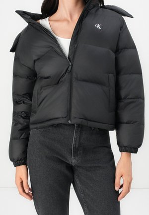 Femme portant une doudoune noire avec une fermeture éclair avant partiellement ouverte, associée à un jean gris foncé et un haut blanc en dessous.