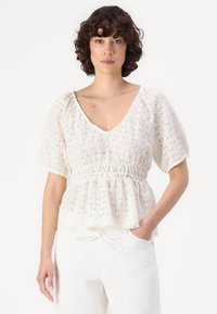 Blouse peplum blanche en dentelle texturée avec un col en V, des manches courtes bouffantes et un détail de taille froncée. Assortie avec un pantalon blanc.
