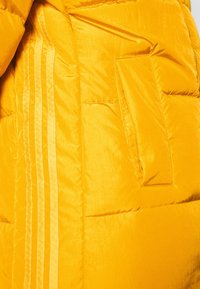 Gul puffjacka med en slät, glänsande yta. Har en dragkedjsficka och tre vertikala ränder längs sidan.