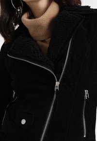 Giacca nera in finta suede con colletto in shearling, frontale con zip e molteplici tasche; presenta hardware argentato e una finitura testurizzata.