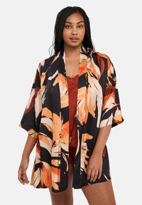 Femme avec des cheveux tressés portant une robe kimono noire ornée de grands imprimés floraux orange et beige sur de la lingerie avec des bordures en dentelle couleur rouille.