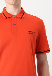 Oranžinė polo marškinė su raukšlėtu apykakle, juodomis juostelėmis ir siuvinėtu "Porsche BOSS" logotipu kairiajame krūtinės pusėje. Dviejų mygtukų užsegimas.
