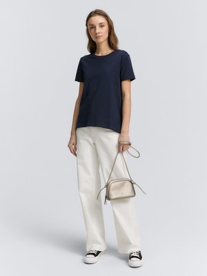 Vrouw staand, gekleed in een marineblauw T-shirt, witte broek met wijde pijpen, zwart-witte sneakers, en een beige kleine crossbodytas vasthoudend aan de bandjes.