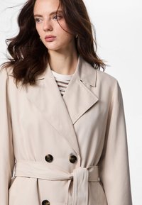 Ung kvinne med bølgete brunt hår som har på seg en beige trenchcoat med belte over en stripete skjorte, ser til siden mot en hvit bakgrunn.