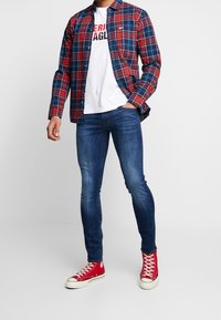 Röd- och blåmönstrad rutig skjorta över en vit t-shirt med svart text, kombinerad med blå jeans och röda sneakers.