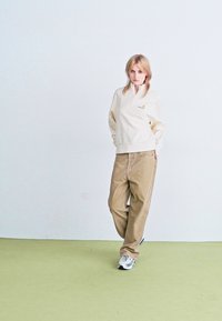 Felpa leggera in pile con zip color crema, polsini a costine e un piccolo logo, abbinata a pantaloni in corduroy beige larghi e scarpe da ginnastica grigie.