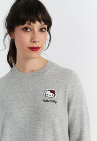 Grijze trui met een ronde hals, zachte textuur en een geborduurd Hello Kitty-logo met een rode strik en de tekst "Hello Kitty".