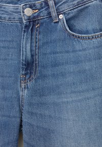 Modré džínové kalhoty s klasickým pětipokládkovým designem. Mají viditelné prošití, kovové knoflíky v pase a mírně vybledlou strukturu.