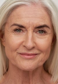 Nærbilde av en moden kvinnes ansikt med sølvhvitt hår, synlige rynker, grønnaktig-grå øyne og en jevn, lys hudtone.