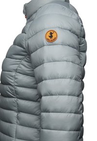 Lichtgrijze pufferjack met een horizontaal gestikt ontwerp. Heeft een ronde oranje patch met de tekst "SAVE THE DUCK" op de linker schouder.