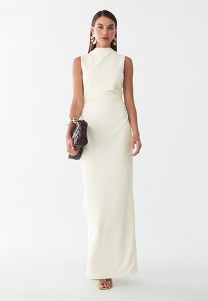 Frau trägt ein ärmelloses weißes Maxikleid, hält eine dunkel geflochtene Clutch und ist mit großen goldenen Ohrringen, Armband und goldenen Sandalen geschmückt.