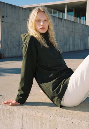 Une femme blonde vêtue d’une veste vert foncé et d’un pantalon blanc s’appuie en arrière contre un muret en béton devant un bâtiment moderne.