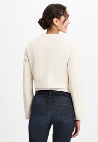 Geknipte crèmekleurige jas met lange mouwen en naaddetails, gecombineerd met donkerblauwe jeans met achterzakken en een platte tailleband.