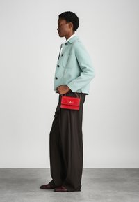 Borsa a mano rossa con effetto coccodrillo e tracolla in argento, indossata accanto a una giacca verde menta, completata da pantaloni marroni su misura.