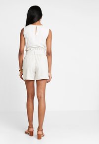 Top bianco senza maniche con un'apertura a chiave sul retro, abbinato a pantaloni corti a righe chiare con vita elastica e cintura in evidenza, indossato con sandali con tacco marroni.