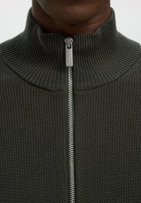 Gros plan d'une personne à la peau foncée portant un pull en tricot côtelé gris foncé avec une fermeture éclair argentée remontée jusqu'au cou.