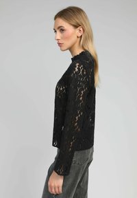 Blouse en dentelle noire avec des manches longues, décolleté festonné, motif floral et texture transparente, assortie à un jean gris.